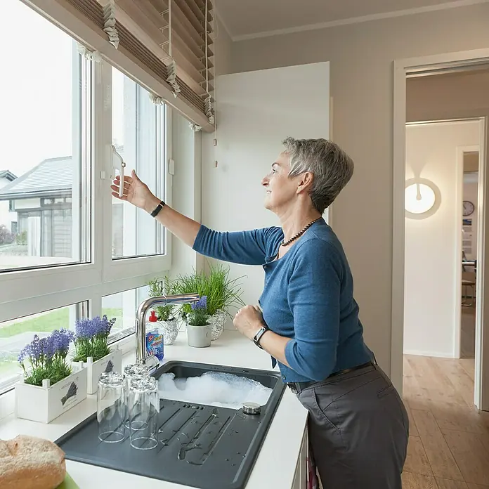 Abus Alarm-Fenstergriff FG 300A - DIN Anschlag: Universell einsetzbar, Weiß, Abschließbar - Frau am Spültisch mit Spülbecken und Abtropfschale, öffnet Fenster und blickt nach draußen. Küche mit Geschirr und Spülmittel.