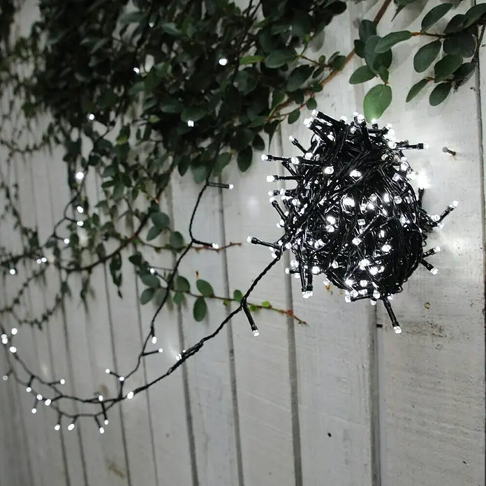Tween Light Guirnalda luminosa LED - Para exterior, Color de luz: Blanco cálido, Número de bombillas: 400 ud. - Guirnalda de luces decorativa con luces blancas, parcialmente enrollada en una pared de madera blanca con plantas verdes.