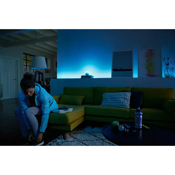 Philips Hue Set de bombillas LED Kit con mando a distancia - E27, Capacidad de atenuación: Intensidad regulable, Blanco cálido, 2.478 lm, 10 W, 3 ud. - Mujer atándose los cordones frente a un sofá verde con iluminación inteligente. La iluminación ambiental crea detalles modernos en la estancia.