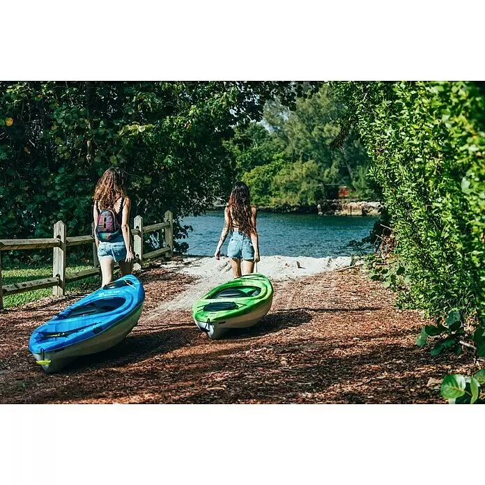 Pelican Kajak Argo 100X - L x B: 305 x 71 cm, Tragkraft: 125 kg, Geeignet für: 1 Person, Blau/Weiß - Zwei Frauen ziehen ein blaues und ein grünes Kajak zum Wasser, bereit für ein Outdoor-Abenteuer.