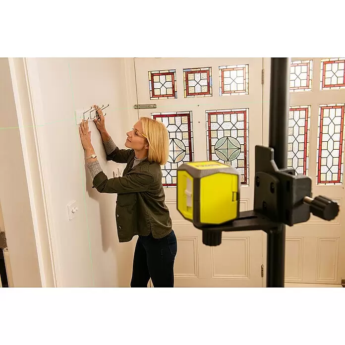 Ryobi Kreuzlinienlaser RBCLLG1 - Max. Arbeitsbereich: 15 m - Gelb-schwarzer Laser-Nivellierer projiziert grüne Linien, die einer Frau beim Anbringen einer Garderobe helfen.