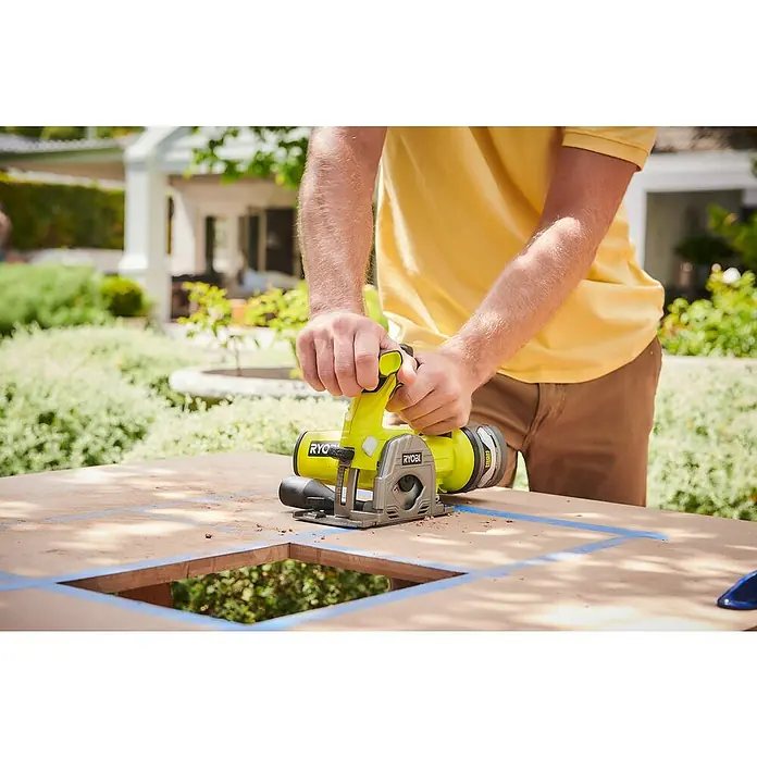 Ryobi ONE+ Akku-Tauchsäge R18MMS-0 - 18 V, Ohne Akku, Leerlaufdrehzahl: 4.250 U/min - Ryobi Akku-Kompaktkreissäge im Einsatz beim Zuschneiden eines Werkstücks im Freien. Das Werkzeug liegt sicher in den Händen des Benutzers.