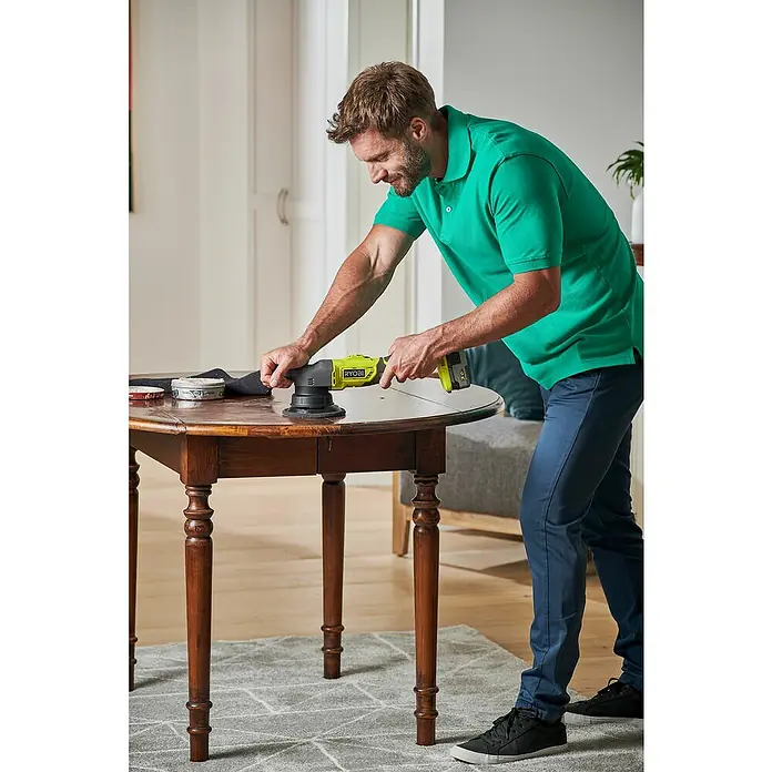 Ryobi ONE+ Akku-Exzenterpolierer R18P-0 - 18 V, Ohne Akku, 125 mm - Ein Mann mit einem grünen Poloshirt schleift einen Holztisch mit einem gelben Ryobi-Schleifer.
