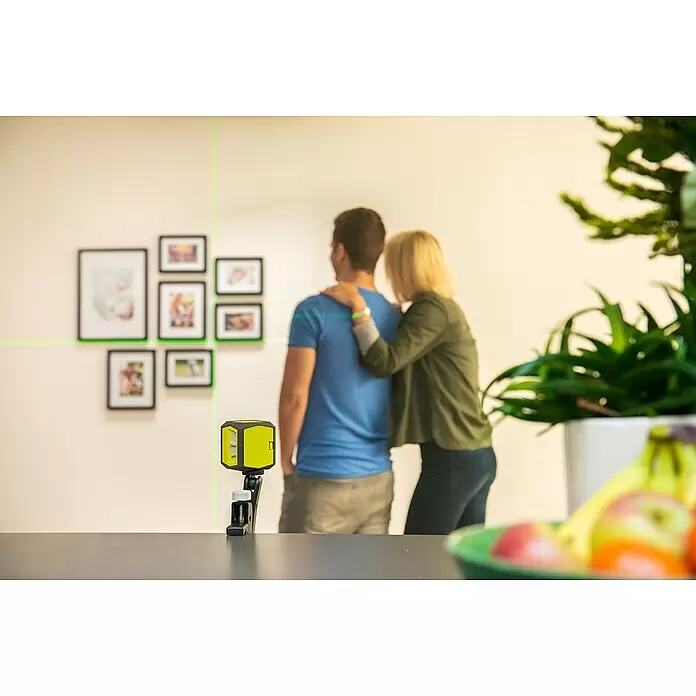 Ryobi Kreuzlinienlaser RBCLLG1 - Max. Arbeitsbereich: 15 m - Gelb-schwarze Laser-Wasserwaage projiziert grüne Linien an eine Wand zum präzisen Ausrichten von Bildern, beobachtet von einem Paar.