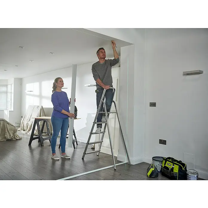 Ryobi Kreuzlinienlaser 360° RB360GLL - Max. Arbeitsbereich: 25 m - Ein Mann auf einer Leiter misst mit einem Laser die Decke, während eine Frau ein Bauteil hält. Werkzeug und Farbe stehen bereit.