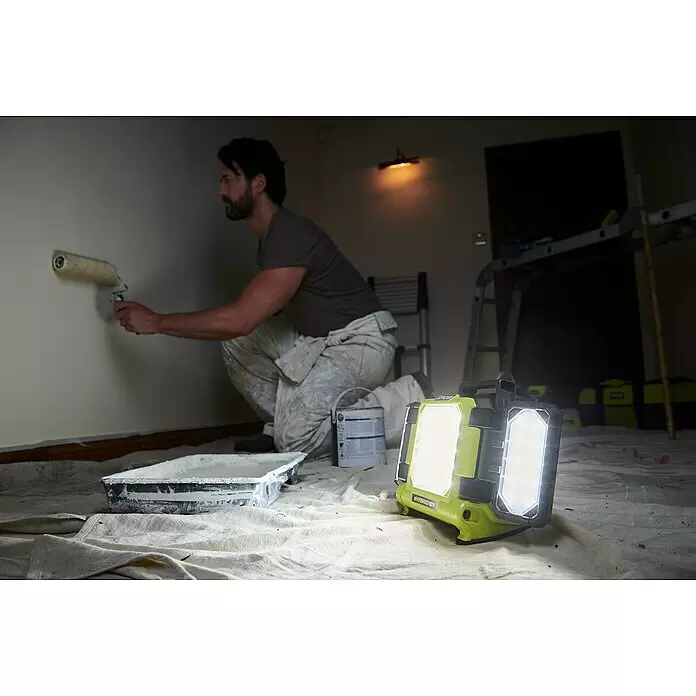 Ryobi ONE+ LED-Akkustrahler RLP18-0 - 18 V, Ohne Akku, 3.000 lm - Eine hellgrüne und schwarze Arbeitsleuchte beleuchtet einen Mann, der in einem dunklen Raum eine Wand streicht.