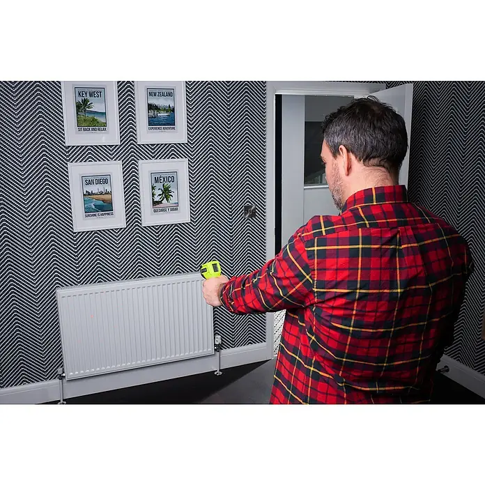 Ryobi Infrarot-Thermometer RBIRT08 - Messbereich: -50 °C bis +500 °C - Ein Mann im rot karierten Hemd benutzt ein Messgerät an einem Heizkörper vor einer Wand mit Bildern und Zickzackmuster.