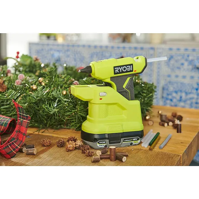 Ryobi ONE+ Akku-Heißklebepistole RGLM18-0 - 18 V, Ohne Akku, Durchmesser Klebepatrone: 7 mm - Nahaufnahme einer RYOBI Akku-Heißklebepistole mit Akku, vor einem Weihnachtskranz und Holzzuschnitten auf einem Tisch.
