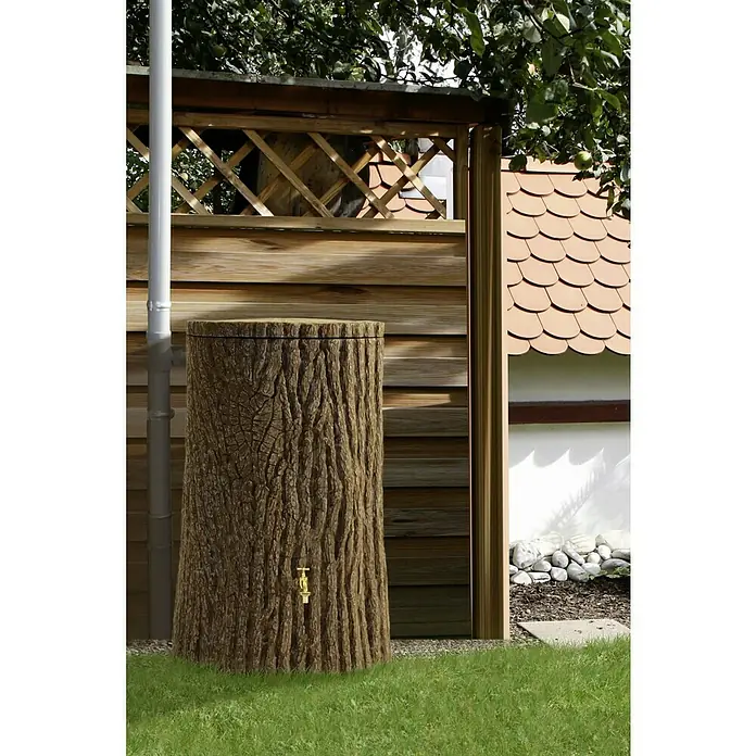 Arves Regenspeicher Evergreen Lite - 300 l, Dunkelbraun - Regentonne in Baumstammoptik mit goldenem Wasserhahn vor Holzzaun und Hauswand. Ein dekoratives und praktisches Element für den Garten.