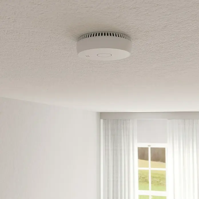 UniTEC Rauchwarnmelder Kriwan/TÜV - Durchmesser: 90 mm, Alarmsignal: 85 dB - Nahaufnahme eines Rauchmelders an einer strukturierten Decke in einem hellen Raum mit Vorhängen vor dem Fenster.