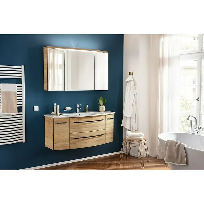 Camargue Swing K Badmöbel-Set BD3 - Gesamtbreite Badmöbel: 120 cm, Berg Eiche, Matt, 3 -tlg. - Modernes Badezimmer mit Waschtischunterschrank in Holzoptik, Spiegelschrank und Handtuchwärmer an einer blau gestrichenen Wand.