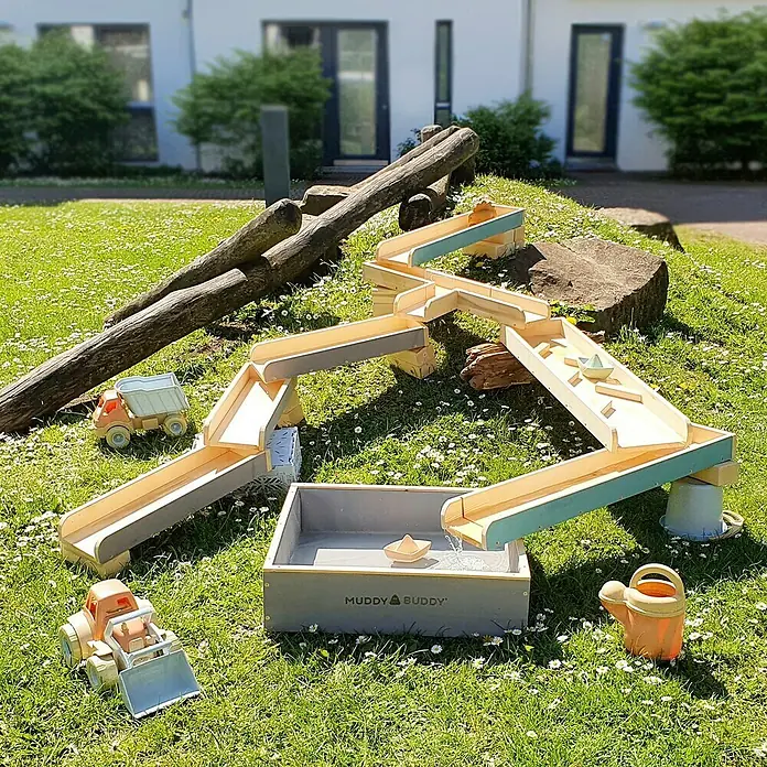 Muddy Buddy Wasserbahn Aqua Traveller - Holz, Natur/Wolkengrau - Holzspielzeug Wasserbahn mit Zubehör, bestehend aus Wasserrad, Wassertisch, Booten, Spielzeugbagger und LKW auf einer grünen Wiese.