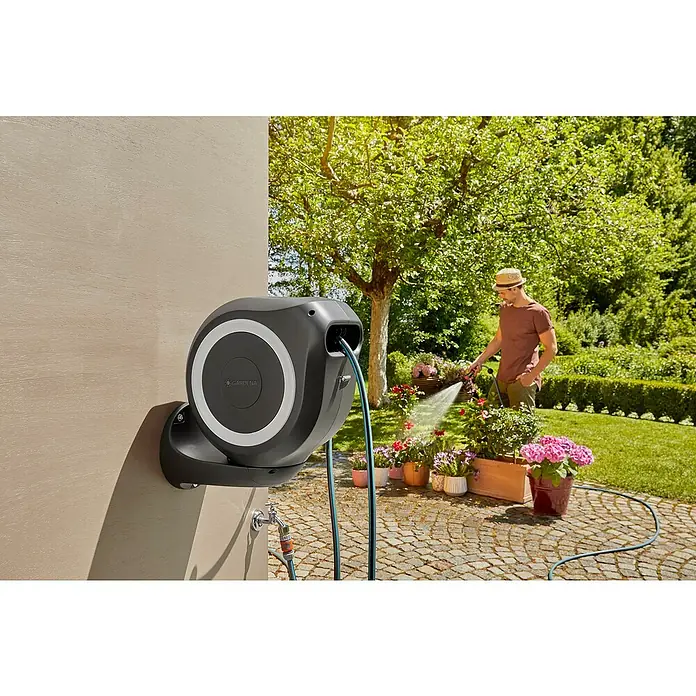 Gardena Wand-Schlauchbox RollUp M - Schlauchlänge: 20 m - Automatische Schlauchbox von Gardena an der Wand, mit Mann im Hintergrund, der Blumen gießt. Ideal für die Gartenbewässerung.