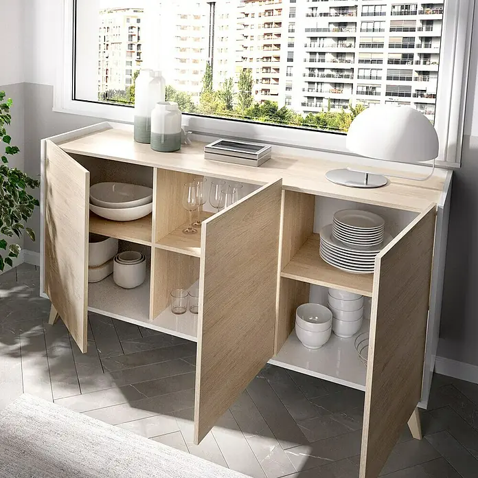 Dekit Aparador Ness - L x An x Al: 43 x 155 x 75 cm, Natural/Blanco - Un moderno aparador de madera clara con puertas abiertas, que muestra platos y vasos. Está situado frente a una ventana con vistas a la ciudad.