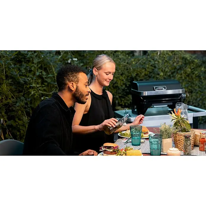 Campingaz Barbacoa de gas Attitude 2GO - Número de quemadores: 1 - Una mujer vierte aceite sobre una hamburguesa en una mesa, con una parrilla negra al fondo.