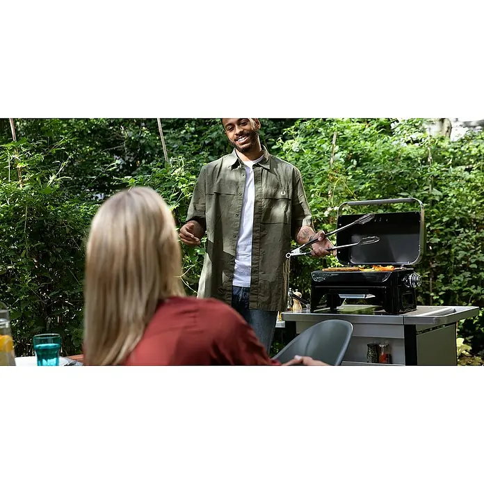 Campingaz Barbacoa de gas Attitude 2GO - Número de quemadores: 1 - Un hombre asa al aire libre en una barbacoa portátil. Utiliza unas tenazas para voltear la comida, mientras sonríe.
