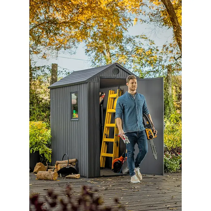 Keter Caseta de jardín Darwin 4x6 - Medidas ext. (An x Pr): 125,8 x 184,5 cm, Plástico, Gris, 2,3 m² - Un hombre sale de un cobertizo de jardín gris con herramientas. Dentro del cobertizo hay una escalera y otras herramientas de jardinería.