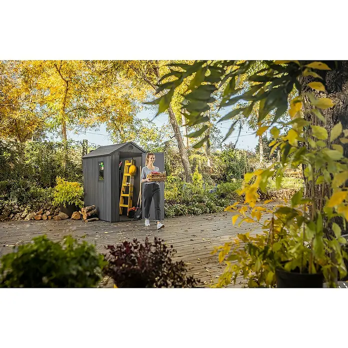 Keter Caseta de jardín Darwin 4x6 - Medidas ext. (An x Pr): 125,8 x 184,5 cm, Plástico, Gris, 2,3 m² - Una mujer sale de un cobertizo de jardín gris, lleno de equipo de jardinería, con una cesta de productos.
