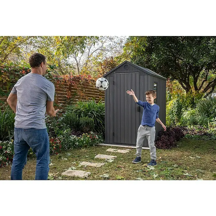 Keter Caseta de jardín Darwin 4x6 - Medidas ext. (An x Pr): 125,8 x 184,5 cm, Plástico, Gris, 2,3 m² - Un padre y su hijo juegan con un balón de fútbol en el jardín delante de un cobertizo gris. El cobertizo ofrece un práctico almacenamiento exterior.
