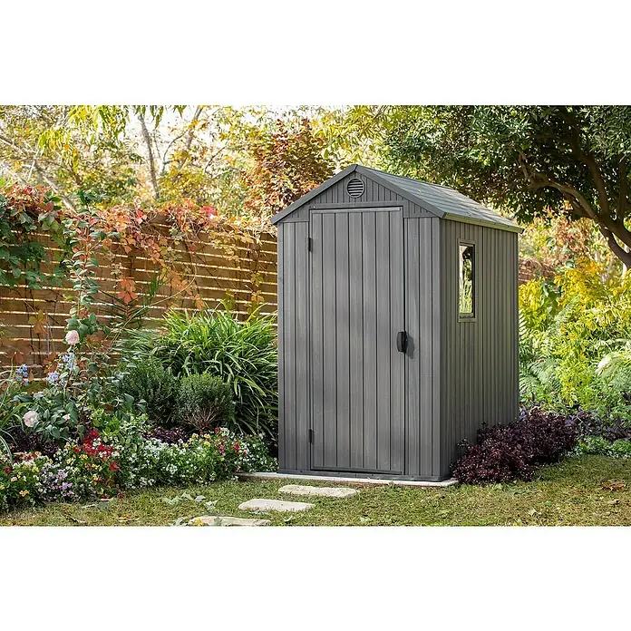Keter Caseta de jardín Darwin 4x6 - Medidas ext. (An x Pr): 125,8 x 184,5 cm, Plástico, Gris, 2,3 m² - Un cobertizo gris con estructura vertical similar a la madera se encuentra en un jardín floreciente. Ofrece un amplio almacenamiento para herramientas y accesorios de jardín.