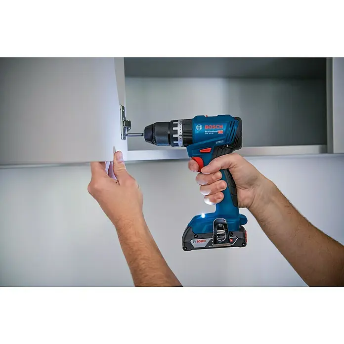 Bosch Professional AMPShare 18V Taladro atornillador percutor de batería GSB 18V-45 - 18 V, 2 baterías, 2 Ah - 4 Ah - Un artesano sujeta un tornillo a un armario blanco con un taladro atornillador azul de Bosch. La herramienta es profesional e inalámbrica.