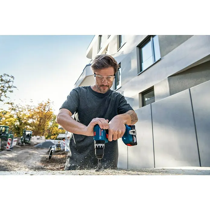 Bosch Professional AMPShare 18V Taladro atornillador percutor de batería GSB 18V-45 - 18 V, 2 baterías, 2 Ah - 4 Ah - Un hombre está perforando agujeros en una tabla de madera con un taladro inalámbrico azul de Bosch. Lleva gafas de seguridad.