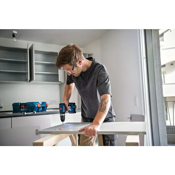 Bosch Professional AMPShare 18V Taladro atornillador percutor de batería GSB 18V-45 - 18 V, 2 baterías, 2 Ah - 4 Ah - Hombre montando una puerta con un taladro inalámbrico Bosch. La caja de herramientas azul de Bosch está al fondo en la encimera de la cocina.