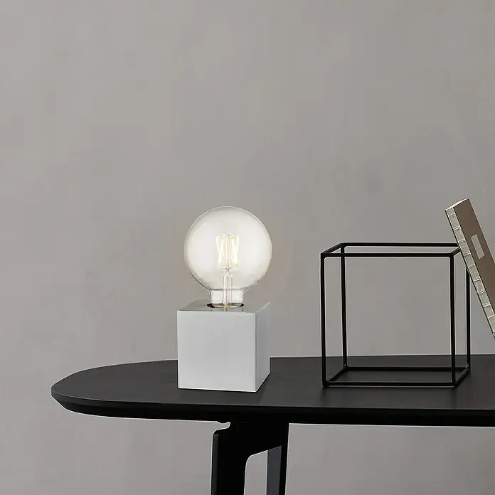 Home Sweet Home Lampenfuß Dry 10 - 10 W, E27, Stahl, Höhe: 8,5 cm - Moderne Tischleuchte mit würfelförmigem Sockel und Glühbirne neben Deko-Objekt auf schwarzem Tisch vor grauer Wand.