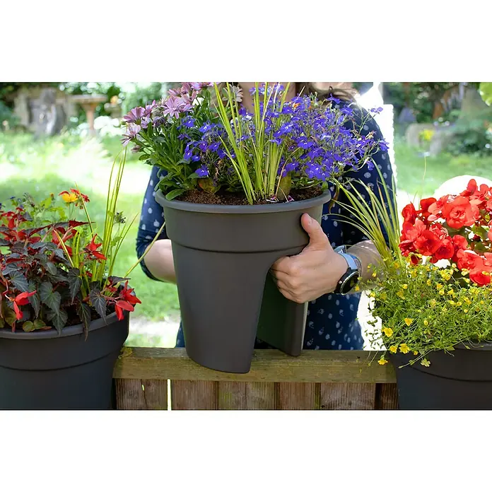 KHW Geländertopf Flowerclip - Außenmaß (Ø x H): 27 x 27,5 cm, Kunststoff, Anthrazit - Nahaufnahme eines Pflanzgefäßes mit bunten Blumen, das von einer Person gehalten wird, neben anderen Pflanzgefäßen auf einem Holzzaun.