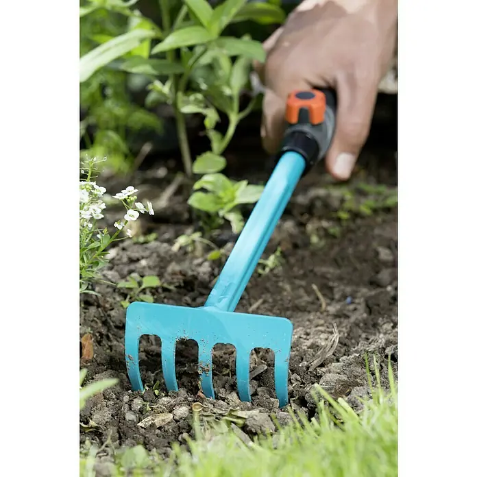 Gardena Combisystem Bügelrechen - Arbeitsbreite: 8,5 cm, Stahl - Nahaufnahme eines hellblauen Rechens, der in der Erde steckt, mit einer Hand am Griff, die das Gartengerät hält.