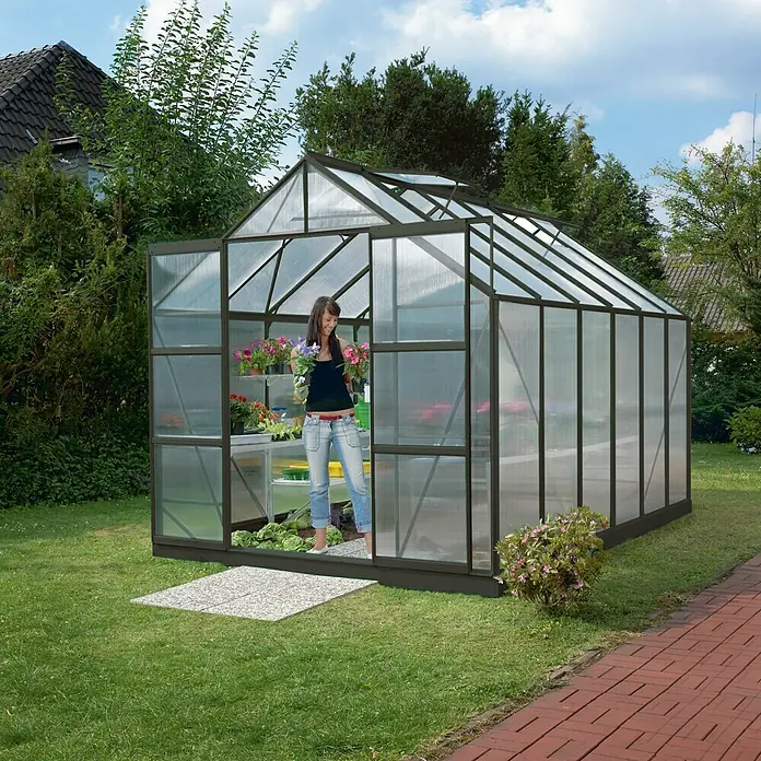 Vitavia Gewächshaus Mars 9900 - Außenmaß inkl. Dachüberstand (B x T): 257 x 383 cm, 6 mm, Schwarz - Eine Frau steht in einem Gewächshaus aus Glas und Metall, umgeben von Pflanzen und Blumen. Der perfekte Ort für Gärtner!