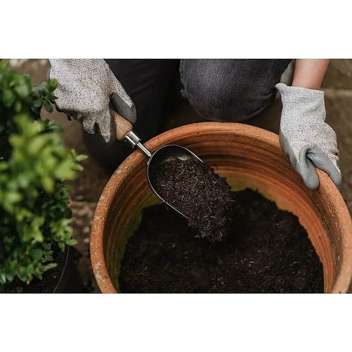 Kent & Stowe Pflanzkelle - 9 cm - Nahaufnahme einer Person, die mit Gartenhandschuhen Erde mit einer Schaufel in einen Terrakotta-Blumentopf füllt, um Pflanzen einzusetzen.