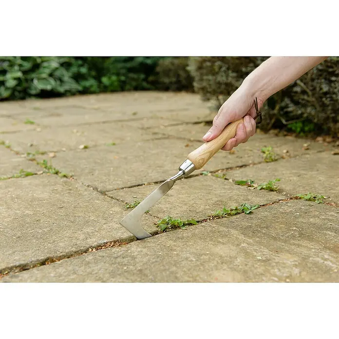 Kent & Stowe Fugenkratzer - Edelstahl - Hand hält einen Fugenkratzer mit Holzgriff, der Unkraut zwischen Pflastersteinen entfernt. Präzises Werkzeug für saubere Gartenwege.