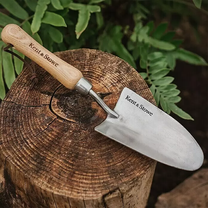 Kent & Stowe Blumenkelle - 8,5 cm - Eine Kent & Stowe Gartenschaufel mit Holzgriff und silberner Kelle ruht auf einem Baumstamm.