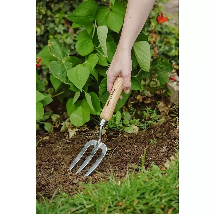 Kent & Stowe Handgabel - 5 cm, 3 - Hand hält eine Kent & Stowe Gartenkralle mit Holzgriff, die in braune Erde zwischen grünen Pflanzen gräbt.
