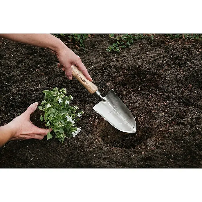Kent & Stowe Blumenkelle - 32 cm - Nahaufnahme einer Person, die eine Kent & Stowe Pflanzkelle benutzt, um eine Blume in dunkle Erde zu pflanzen.