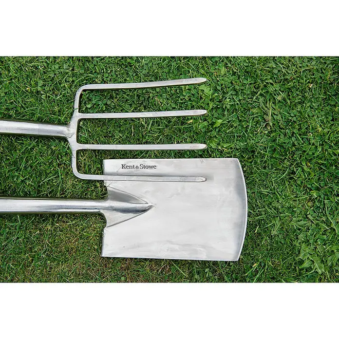 Kent & Stowe Spatengabel - Maße Gabel: 300 x 170 mm, Anzahl Zinken: 4, Langstiel - Nahaufnahme einer glänzenden Kent & Stowe Gartengabel und eines Spatens auf grüner Wiese, die hochwertige Verarbeitung betonend.