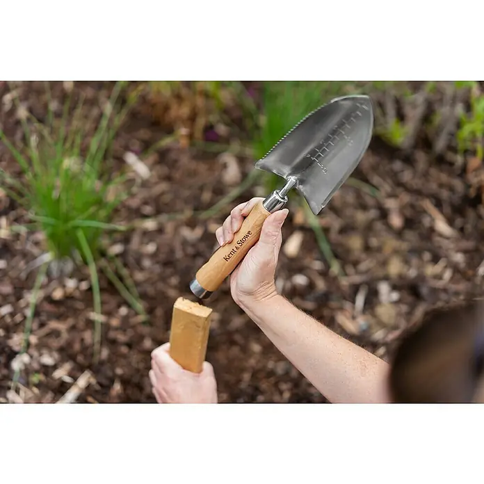 Kent & Stowe Blumenkelle - 32 cm - Nahaufnahme einer Hand, die eine Kent & Stowe Pflanzkelle mit Holzgriff über einem Stück Holz im Garten hält.