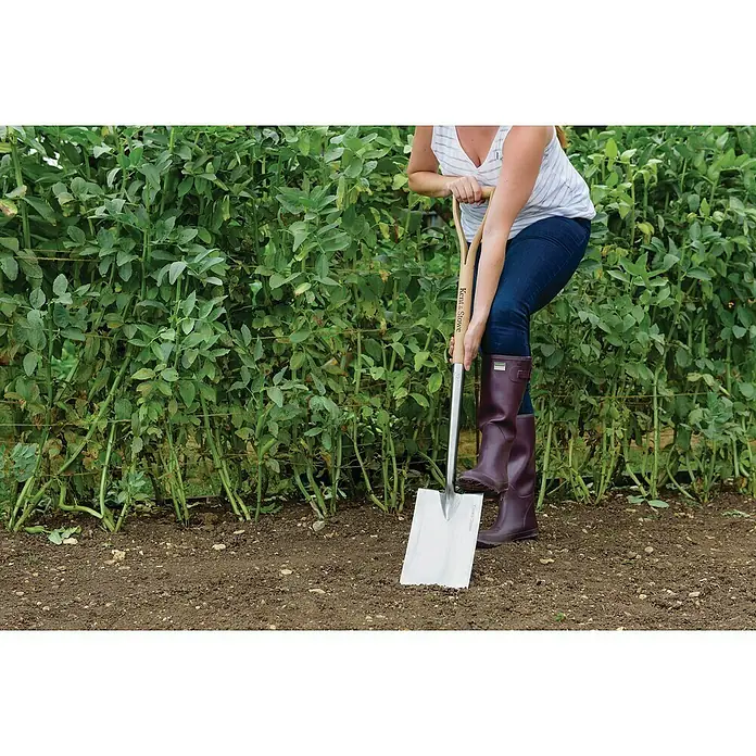 Kent & Stowe Gärtnerspaten - Arbeitsbreite: 18 cm, Langstiel - Nahaufnahme einer Frau, die mit einem Kent & Stowe Spaten in einem Gartenbeet arbeitet, ideal für Gartenarbeiten.