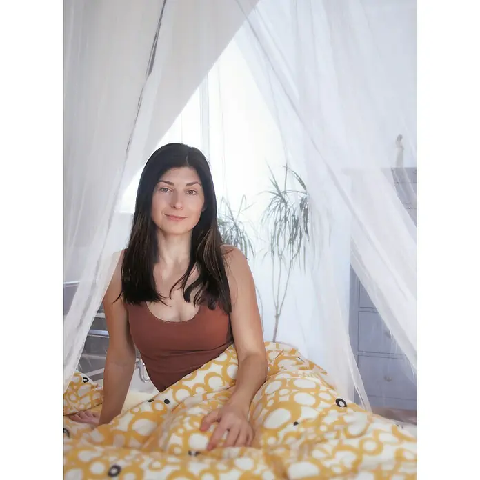 Schellenberg Mosquito Net Mosquitera Plus - An x Al: 250 x 120 cm, Montaje de la mosquitera: Colgar - Mujer sentada en la cama bajo un mosquitero, envuelta en un edredón amarillo con diseño de círculos y con una camiseta de tirantes marrón.