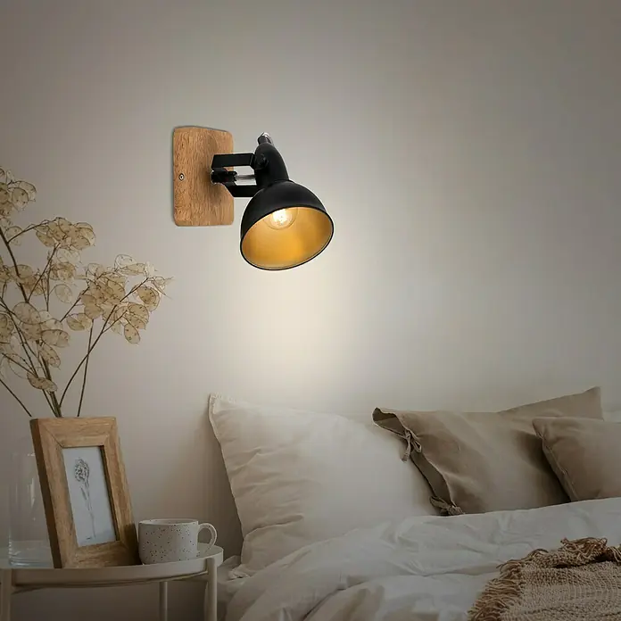 Briloner Barnim Aplique de pared redondo - 25 W, Ø x Al: 11 x 15,5 cm, Negro/Marrón, Negro/Dorado, E14 - Elegante lámpara de pared con pantalla negra e interior dorado, montada sobre una base de madera, que emite una luz cálida en un dormitorio.