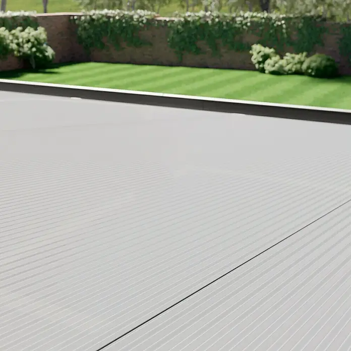 Gardendreams Terrassenüberdachung Comfort - B x T: 604 x 300 cm, Anthrazitgrau, Polycarbonat, Transparent - Eine Nahaufnahme von  einem gerippten, grauen Terrassendielenboden mit einem Rasen und einer Ziegelmauer mit Pflanzen im Hintergrund.