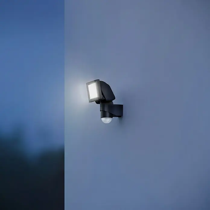 Steinel LED-Sensor-Strahler LS 150 S schwarz - 14,7 W, L x B x H: 17 x 15,5 x 21,5 cm, Schwarz, IP44 - Außenleuchte mit Bewegungsmelder an einer blauen Wand, die nachts für Sicherheit und Komfort sorgt.