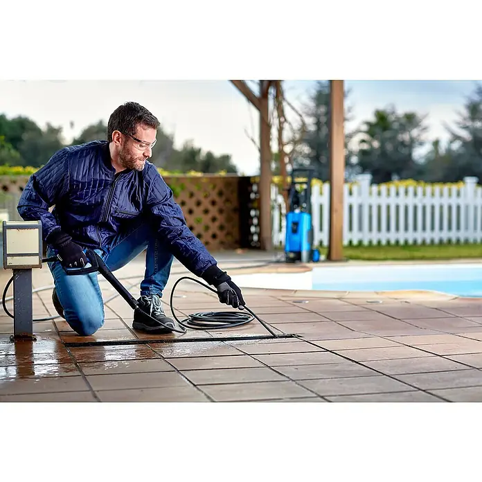 Nilfisk Juego para limpieza de tuberías - 15 m, Específico para: Limpiadoras de alta presión Nilfisk - Hombre limpiando baldosas de patio con una hidrolimpiadora. El aparato azul está al fondo junto a la piscina.