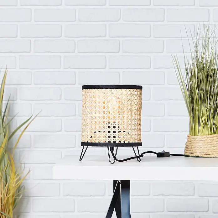 Brilliant Wiley Tischleuchte - 60 W, Schwarz, E27 - Nahaufnahme einer stilvollen Tischlampe mit Rattanschirm und schwarzem Metallfuß, platziert auf einem weißen Tisch vor einer Backsteinwand.