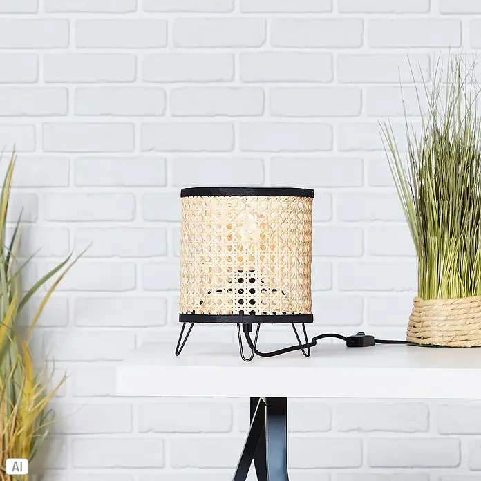 Brilliant Wiley Tischleuchte - 60 W, Schwarz, E27 - Nahaufnahme einer stilvollen Tischlampe mit Rattanschirm und schwarzem Metallfuß, platziert auf einem weißen Tisch vor einer Backsteinwand.