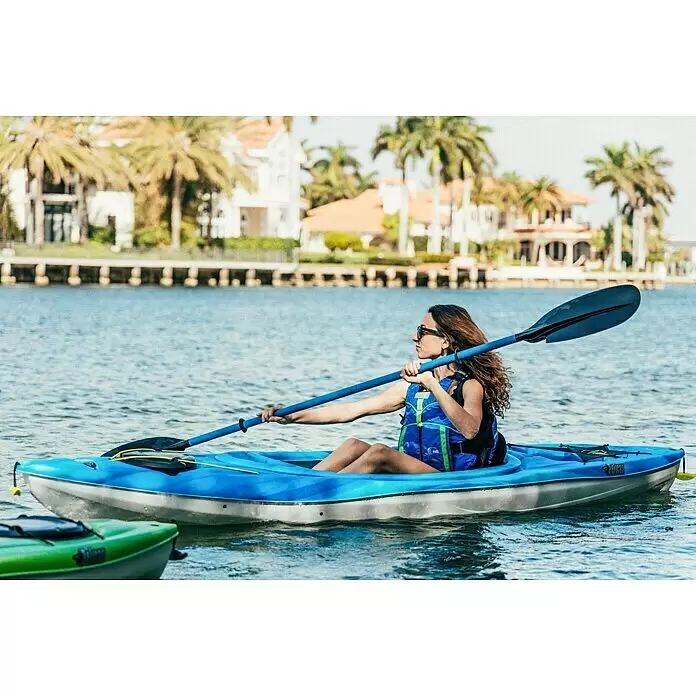 Pelican Kajak Argo 100X - L x B: 305 x 71 cm, Tragkraft: 125 kg, Geeignet für: 1 Person, Blau/Weiß - Frau im blauen Kajak mit Paddel und Schwimmweste auf dem Wasser nahe Häusern.