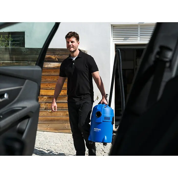 Nilfisk Aspirador en seco y húmedo eléctrico Buddy II 18 - 1.200 W, Volumen de recipiente: 18 l, Con bolsa - Hombre llevando un aspirador azul Nilfisk Buddy II 18 para sólidos y líquidos fuera de un coche. Práctico y potente.
