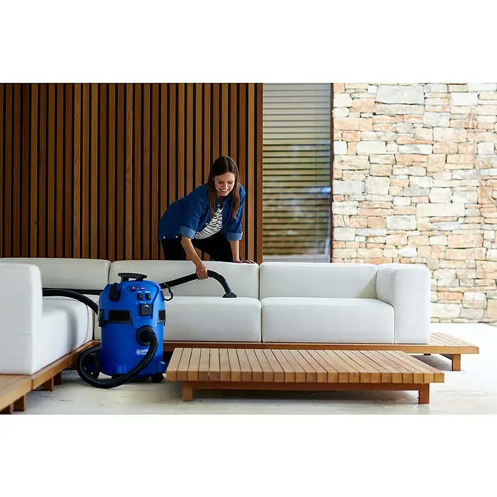 Nilfisk Elektro-Nass- & Trockensauger Multi II 30 T EU - 1.400 W, Behältervolumen: 30 l, Halbautomatisch - Eine Frau reinigt eine weiße Couch mit einem blauen Nilfisk-Reinigungssauger. Der Sauger ist auf einem Holzpodest neben einem passenden Tisch platziert.
