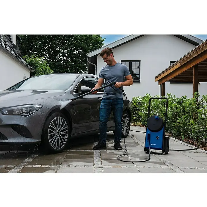 Nilfisk Hidrolimpiadora de alta presión CORE 150 PC - 150 bar, 2.000 W, Capacidad de bombeo: 468 l/h - Un hombre está usando una hidrolimpiadora Nilfisk de alta presión para limpiar un coche gris con una casa al fondo.